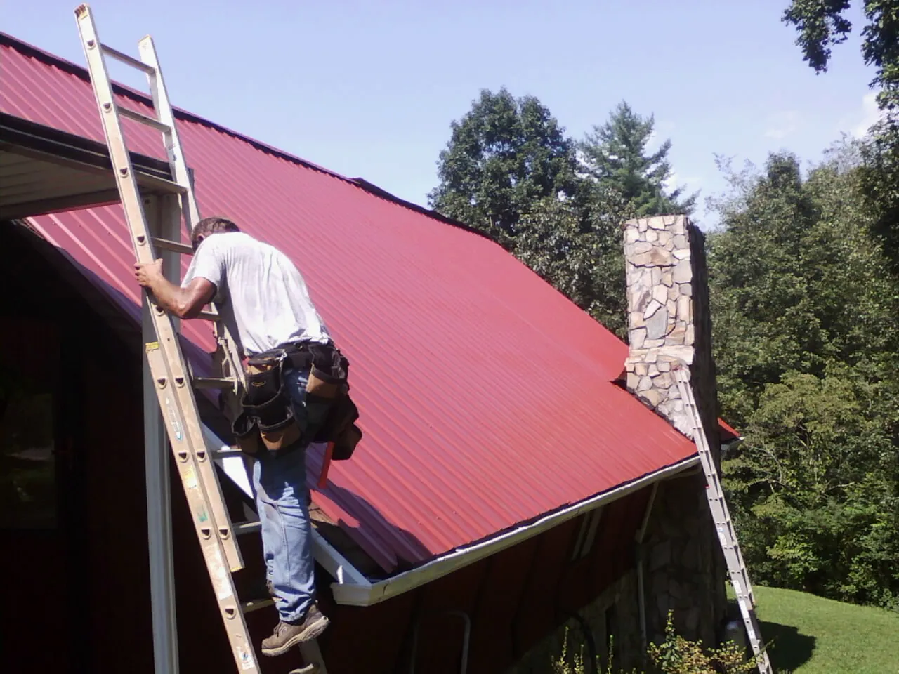 Roofer on a ladder beside a red metal roof and stone chimney — Soffit & Fascia Repair by Roofing Brothers in Pass Christian